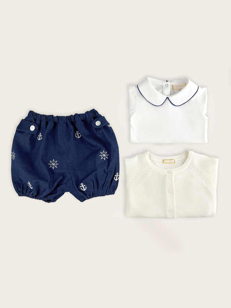 Baby boy bloomer in a blue denim with white nautical anchors and ship's wheels paired with a winter white merino wool cardigan and white body suit with blue piping on the collar.