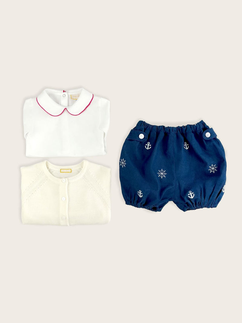 Baby boy bloomer in a blue denim with white nautical anchors and ship's wheels paired with a winter white merino wool cardigan and white body suit with red piping on the collar.